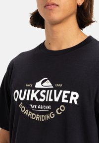 Camiseta de algodón negra con un audaz logo de Quiksilver en blanco y dorado con un diseño de ola, cuello redondo y mangas cortas.