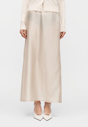 Ženska oblečena v dolgo, bež satensko krilo z belimi čevlji z ostrim prstom in zapestnicami iz biserov okoli gležnjev, stoji pred enobarvnim ozadjem.