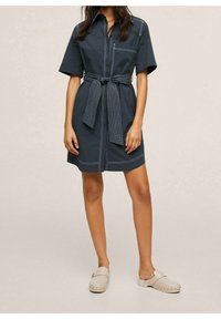 Robe courte à manches courtes bleu marine foncé avec une fermeture éclair avant, une taille ceinturée, une poche poitrine et des surpiqûres blanches contrastantes sur le tissu. Portée avec des sabots clairs.