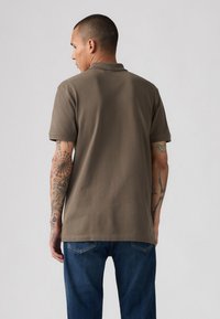 Bruin polo shirt met korte mouwen, eenvoudige design en een zachte textuur, gecombineerd met blauwe denim jeans, weergegeven vanuit de achterkant.