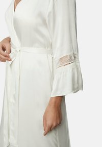 Robe en satin blanc avec une taille nouée et des manches évasées. La dentelle au bord des manches ajoute une touche délicate à la texture lisse.