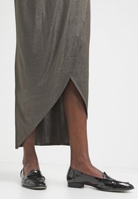 Lång, metallisk olivgrön kjol med en sidosprund, matchad med glänsande svarta loafers. Kjolen har en slät textur och en draperad design.