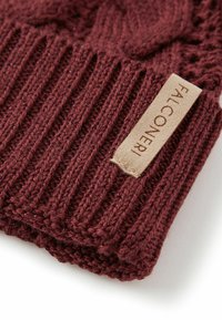 Bordowa dzianinowa czapka typu beanie z żeberkowanym wzorem i beżową metką z wytłoczonym napisem. Tekstura jest miękka z lekko błyszczącym wykończeniem.