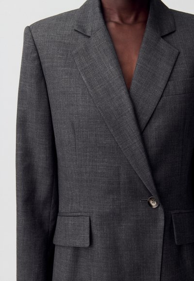 Blazer de lino gris con solapa muesca, cierre de un solo botón y dos bolsillos frontales. Textura suave con un patrón sutil.