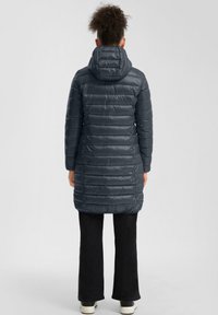 Tamno siva подстavljena jakna с капуљачом, са хоризонталним кутираном секцијама, дужине до колена и глатком, сјајном текстуром. Црне панталоне и обућа.