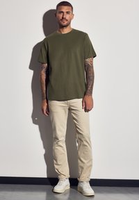 Camiseta de algodón verde oliva de mangas cortas, combinada con jeans beige claros. El modelo lleva zapatillas blancas. El fondo es liso.