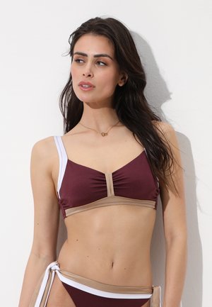 Donna con lunghi capelli scuri che indossa un bikini a due pezzi marrone scuro, beige e bianco, in piedi davanti a uno sfondo bianco uniforme.