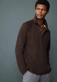 Camisa polo de manga larga marrón con cuello alzado, que presenta un cierre de botón y una tela texturizada. Combinada con pantalones gris claro.