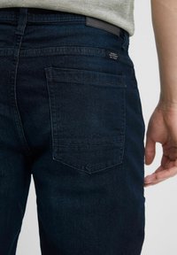 Tumedat sinised denimpüksid sisaldavad tagataskut, millel on peene mägimustri heegeldus. Kudum on sile ja tekstuuriga pinnaga.