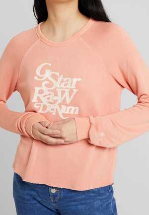 Vrouw draagt een perzikkleurige sweater met ronde hals en de tekst "G Star Raw Denim", haar handen samengevouwen ter hoogte van de taille, gecombineerd met een blauwe spijkerbroek.