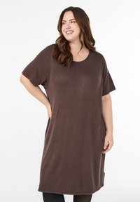 Smaidīga sieviete ar gariem brūniem matiem valkā vaļēju, īsroku brūnu kleitu, stāvot ar roku uz gurna pret vienkrāsainu baltu fonu.