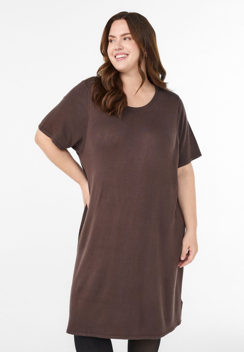 Smaidīga sieviete ar gariem brūniem matiem valkā vaļēju, īsroku brūnu kleitu, stāvot ar roku uz gurna pret vienkrāsainu baltu fonu.