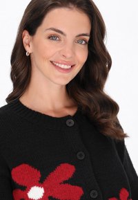 Cardigan lavorato a maglia nero con un design floreale rosso e un centro bianco. Ha un colletto rotondo e bottoni neri sul davanti.