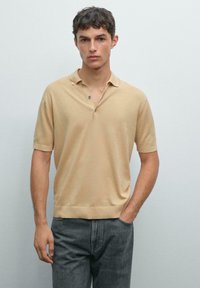 Giovane uomo che indossa una polo beige a maniche corte e jeans grigio scuro, in piedi con una mano in tasca su uno sfondo neutro.