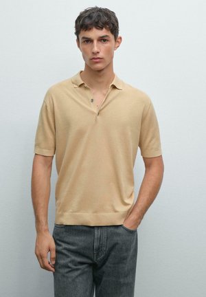 Ung mand iført en beige kortærmet polo-shirt og mørk grå jeans, stående med den ene hånd i lommen mod en ensfarvet baggrund.