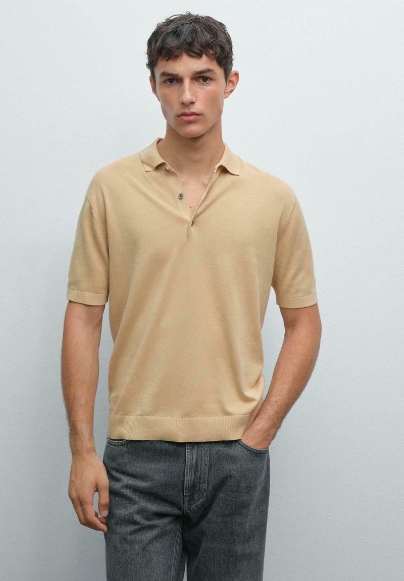 Giovane uomo che indossa una polo beige a maniche corte e jeans grigio scuro, in piedi con una mano in tasca su uno sfondo neutro.