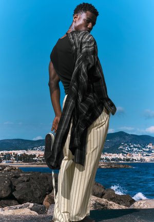 Top negru fără mîneci stratificat cu o cămașă în carouri negre și gri, pantaloni crem cu dungi, ținând o sneaker, pe fundalul unui cer albastru și peisaj de coastă.
