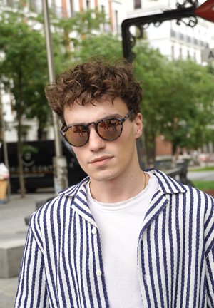 Round sunglasses with dark tinted lenses in a speckled navy and gray frame, paired with a striped white and blue button-up shirt.