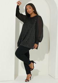 Svart paljett-tunika med genomskinliga, långa ärmar; parat med figurnära svarta byxor och svarta högklackade sandaler med remmar; slät textur.