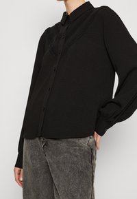 Black long-sleeve blouse with a button-down front, textured fabric, and decorative lace detailing along the chest and shoulders.