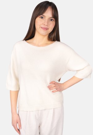 Junge Frau mit langen braunen Haaren, die einen locker sitzenden weißen Pullover und helle Hosen trägt, steht mit einer Hand in der Hüfte vor einem weißen Hintergrund.