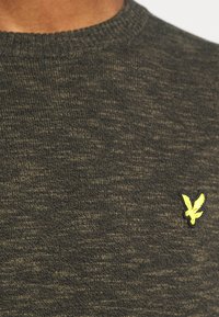 Tummanvihreä neulottu neulepaita, jossa on pyöreä pääntie, teksturoitu kangas ja pieni keltainen kirjailtu kotkalogo vasemman rinnan alueella.