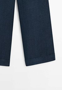 Ourlets pliés bleu foncé de pantalons à jambes droites sur fond blanc, montrant la texture du tissu et les détails des coutures.