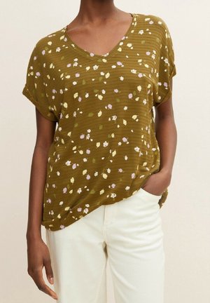 Olivengrøn V-hals bluse med korte ærmer og lille blomstermønster i hvid, lilla og grøn, båret med hvide bukser.
