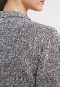 Gros plan d'une personne portant un blazer gris texturé, montrant l'épaule arrière et une partie du cou et du visage de profil.