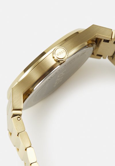 Montre en acier inoxydable doré avec un boîtier octagonal, une finition texturée et une couronne proéminente affichant le logo "BOSS". Les maillons du bracelet sont également dorés.