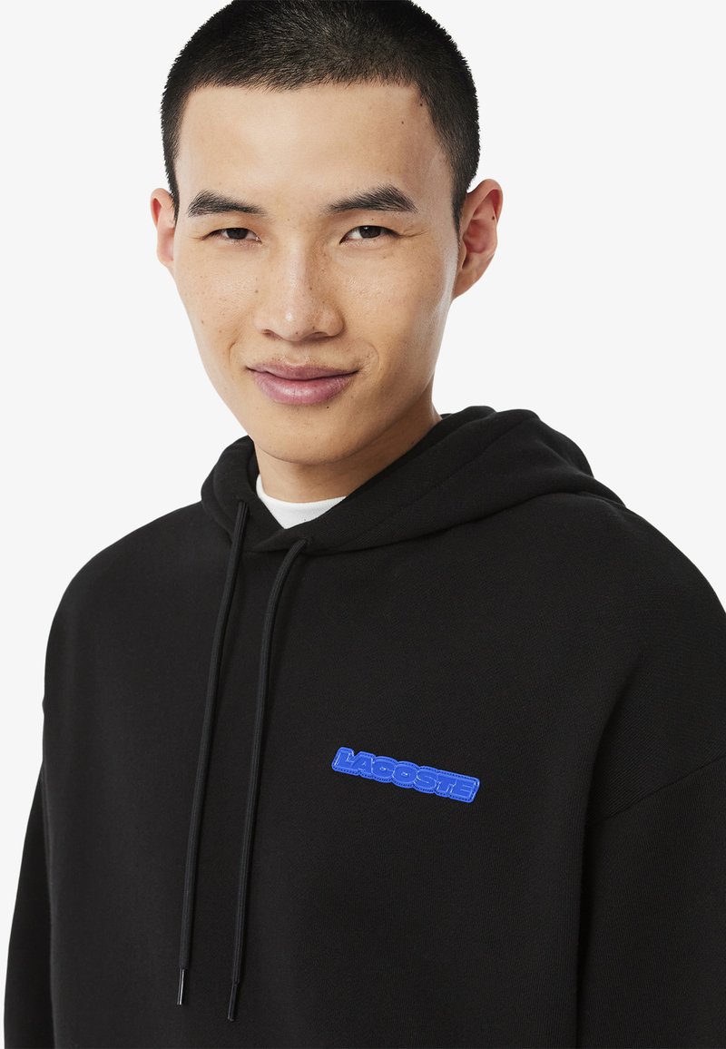 Felpa nera con cappuccio e coulisse, con un'etichetta logo blu Lacoste sul davanti. Tessuto morbido, vestibilità rilassata e design minimalista.