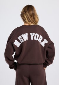 Woman with long hair wearing a brown sweatshirt and pants, sweatshirt back displays large white text "NEW YORK."