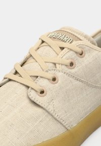 Gros plan sur une basket en tissu beige avec des lacets assortis, des œillets en métal et une semelle en caoutchouc marron clair, montrant les détails de couture et l'étiquette Apple Yard sur la languette.