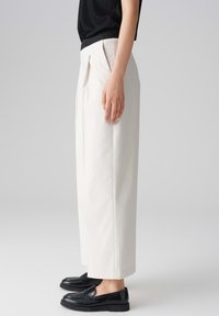 Pantaloni bianchi a pieghe con una vestibilità ampia, realizzati in tessuto testurizzato, abbinati a mocassini neri. La vista laterale mette in mostra la silhouette sartoriale.