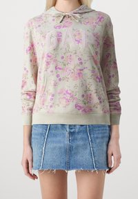 Donna che indossa un hoodie floreale color crema con il testo "GAP" e una mini gonna di jeans blu con orlo sfrangiato, in piedi contro uno sfondo bianco.