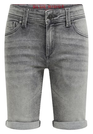 Młoda osoba stoi z jedną ręką w kieszeni, ubrana w pomarańczowy t-shirt w wzory, szare podwinięte szorty jeansowe, białe wysokie skarpetki oraz białe tenisówki.