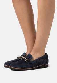 Mörkblå mocka loafers med guldfärgad detalj, platt sula och öppen baksida, som visar upp en slät textur och elegant form.