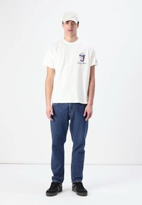Hvit bomull t-skjorte med grafisk popcorn-design og "Tommy Jeans"-tekst, kombinert med blå denimjeans og svarte joggesko. Modellen har på seg en lys caps.