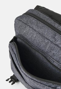 Mochila de tela gris con un acabado texturizado, que cuenta con un compartimento con cremallera y un panel trasero acolchado para mayor comodidad. Incluye correas ajustables.