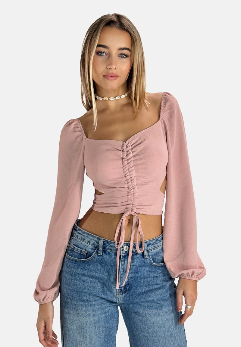 Crop top a maniche lunghe di colore rosa chiaro con dettagli arricciati davanti e chiusura a fiocco, con aperture laterali. Abbinato a jeans blu a vita alta.
