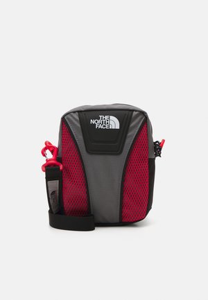 Petit sac bandoulière gris et rouge avec sangle noire réglable et logo blanc The North Face sur le panneau avant.