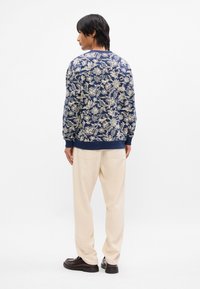 Σαλοπέτα ναυτικού τύπου με λουλουδάτο μοτίβο, φτιαγμένη από υφασμάτινη υφή, με ριγωτό λαιμόκοψι και μανίκια, συνδυασμένη με ανοιχτό μπεζ παντελόνι και μαύρα παπούτσια.