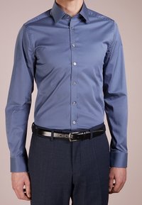 Lichtblauw, overhemd met lange mouwen, een puntkraag, een middenstaande knoopsluiting en zilverkleurige knopen. Draagt bijpassende donkere broek en een zwarte riem.