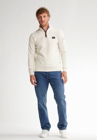 Pulover cu fermoar de un sfert, de culoare crem deschis, cu un fermoar maro și o mică etichetă cu logo, purtat cu blugi albaștri și adidași albi.