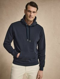 Mörkblå hoodies i mjukt material, med kängurufickor och snörningar. Modellen bär ljusa byxor.