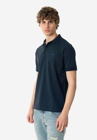 Polo shirt blu navy con colletto classico, maniche corte e un piccolo logo blu sul lato sinistro del petto. Tessuto strutturato, forma aderente e leggero.