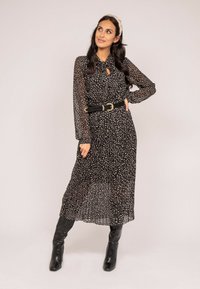 Robe noire à manches longues avec un col noué et des taches blanches. Elle présente une jupe plissée et une large ceinture noire avec une boucle dorée.