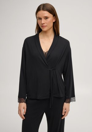 Vrouw die een zwarte wikkelbadjas met kanten manchetten draagt en een bijpassende top met kanten rand daaronder, tegen een effen achtergrond.