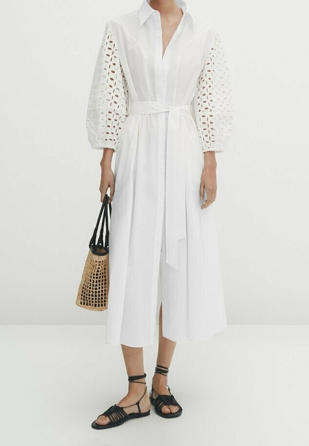 Femme portant une robe midi blanche avec des manches bouffantes à œillets, tenant un sac à main tressé couleur cuir clair, et portant des sandales plates noires à lanières.