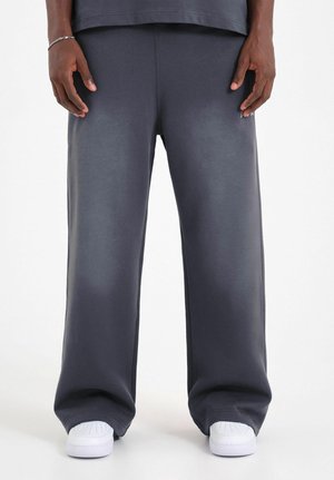 Persona che indossa pantaloni tuta grigio scuro larghi e scarpe da ginnastica bianche, in piedi con le mani rilassate ai lati, su sfondo bianco.